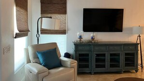 Ein Wohnzimmer mit einem beigefarbenen Sofa, einem an der Wand montierten Fernseher, einem Schrank mit Glastüren und einer Stehlampe.