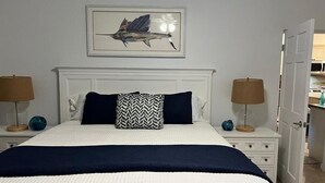 Ein ordentlich bezogenes Bett mit einer weißen und dunkelblauen Tagesdecke, einem Zierkissen und einem gerahmten Kunstwerk darüber. Zwei Nachttische mit Lampen und Dekorationselementen sind vorhanden.