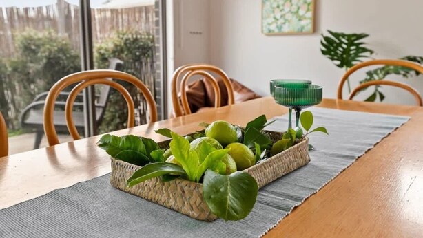 Una mesa de comedor con una cesta tejida que contiene hojas verdes y un objeto verde, sillas de madera y vistas a través de una ventana.