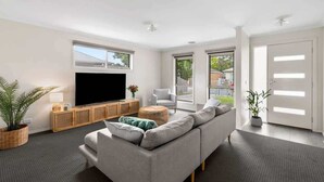 Una sala de estar moderna con un televisor de pantalla plana grande, un sofá gris, un mueble de madera y un ventanal amplio.