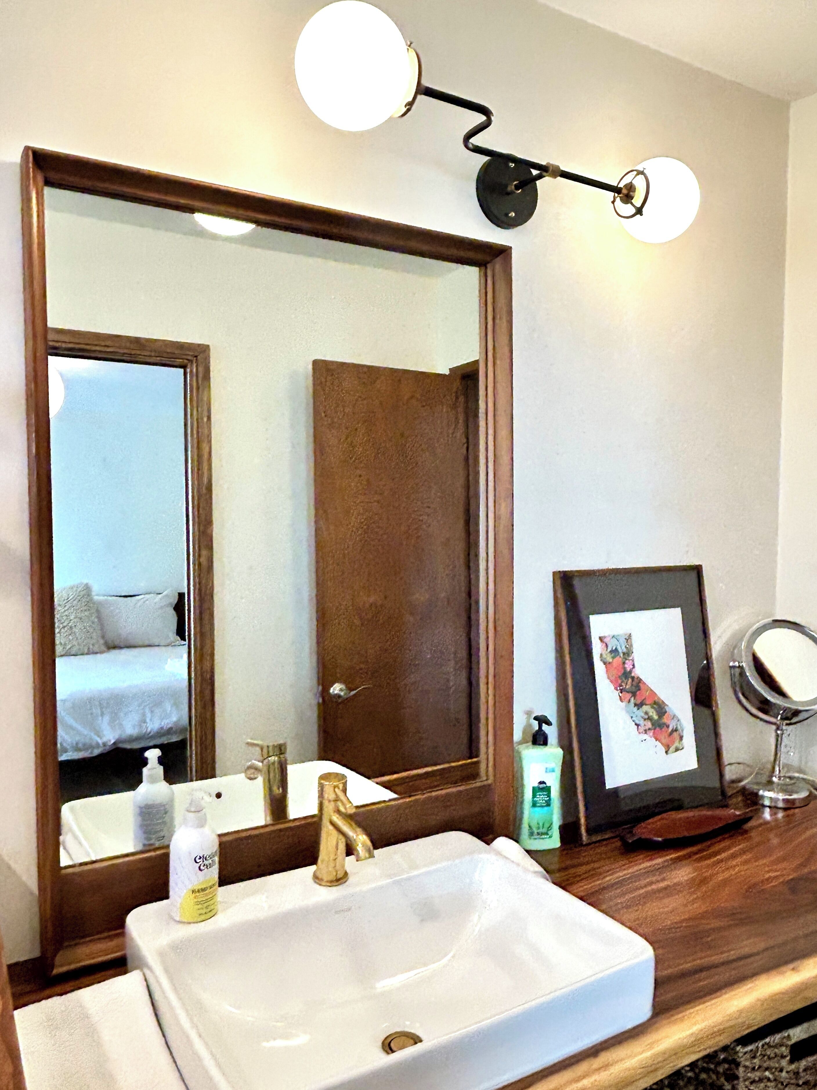 A bathroom with a large mirror, a white sink, a framed picture, and a wall-mounted light fixture.