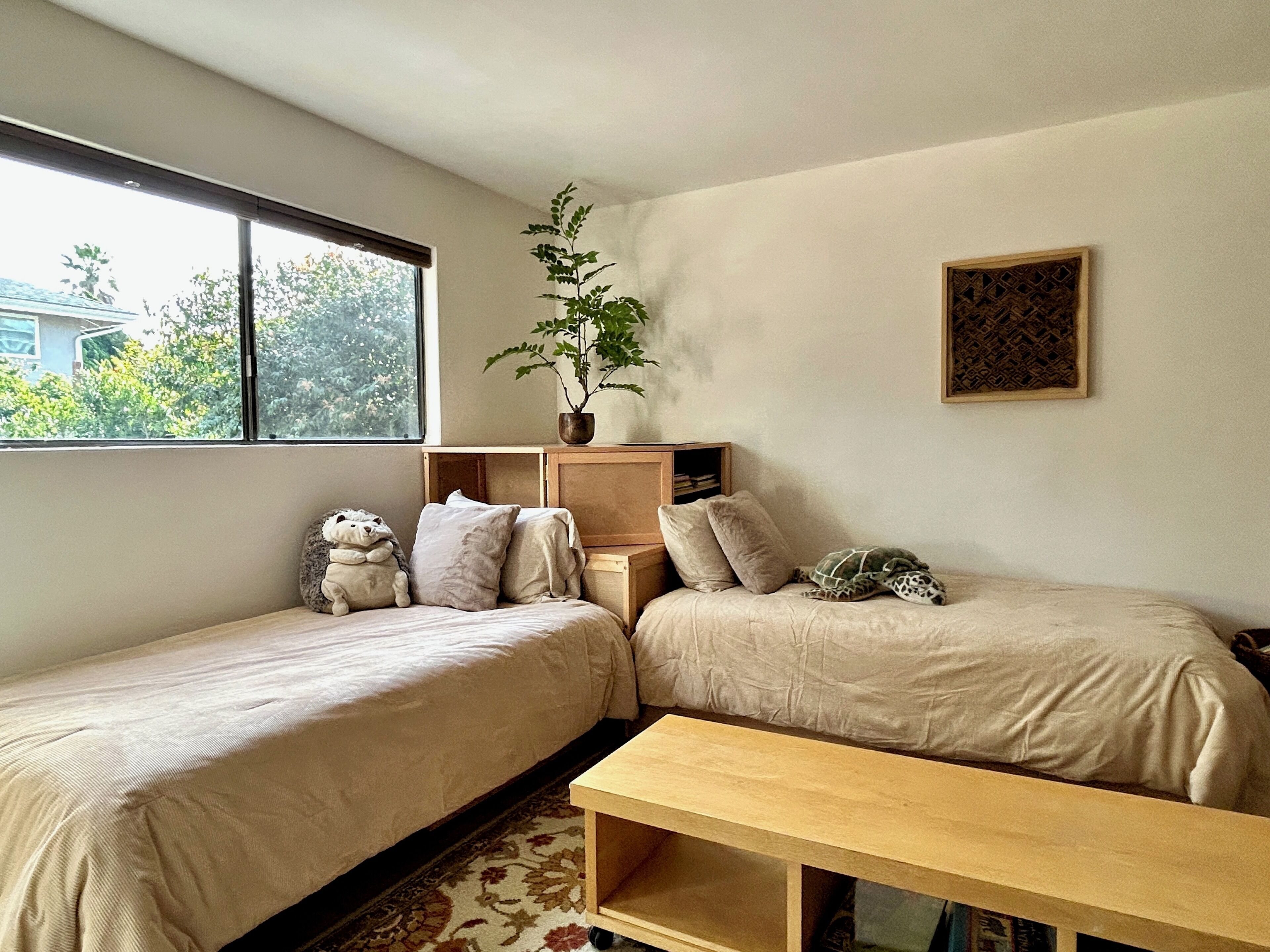 Une chambre avec deux lits simples, une table de chevet en bois, une petite table, une plante en pot et un tableau encadré au mur.