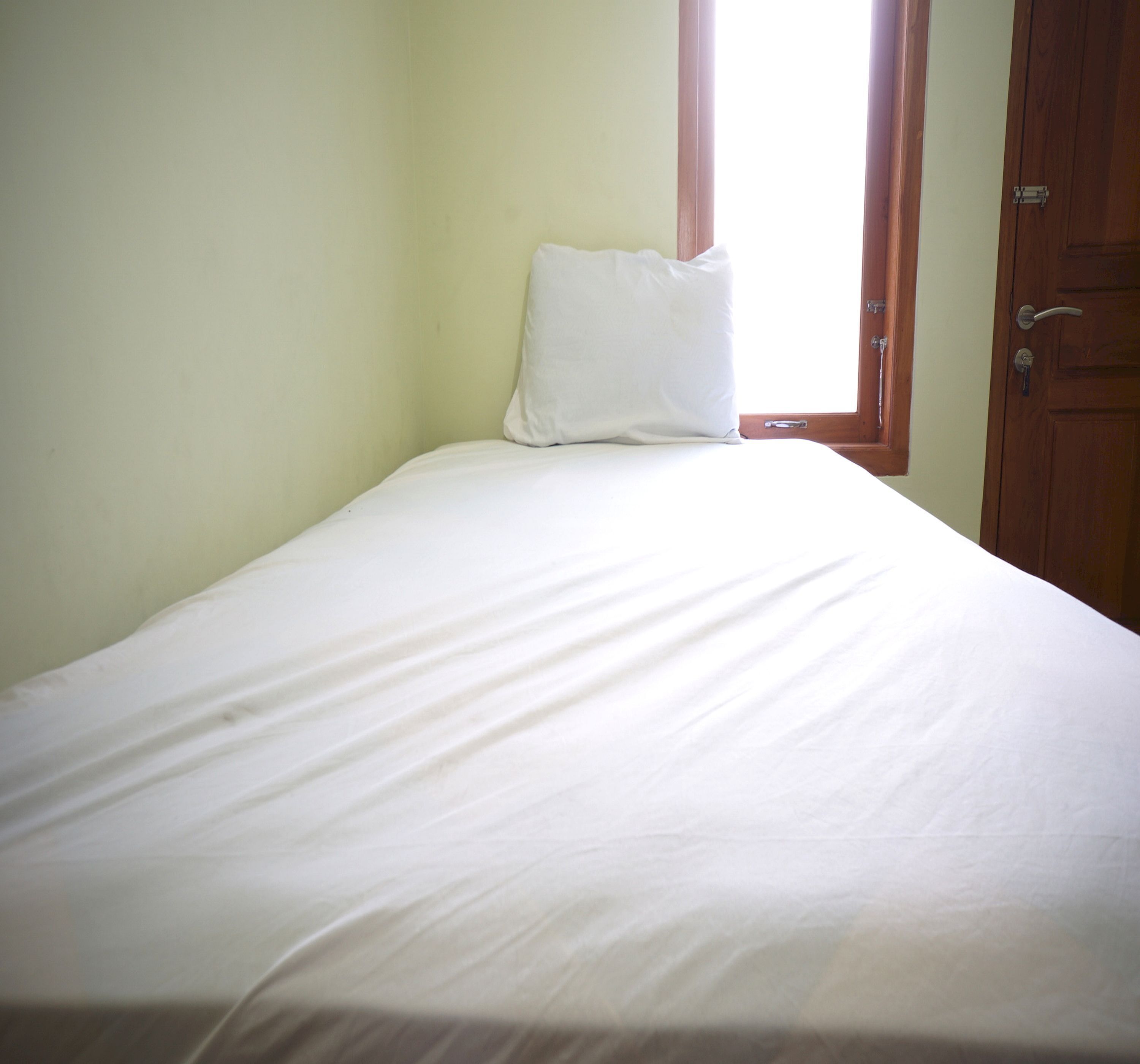 A single bed with a white mattress and pillow in a room with light green walls and a wooden door.