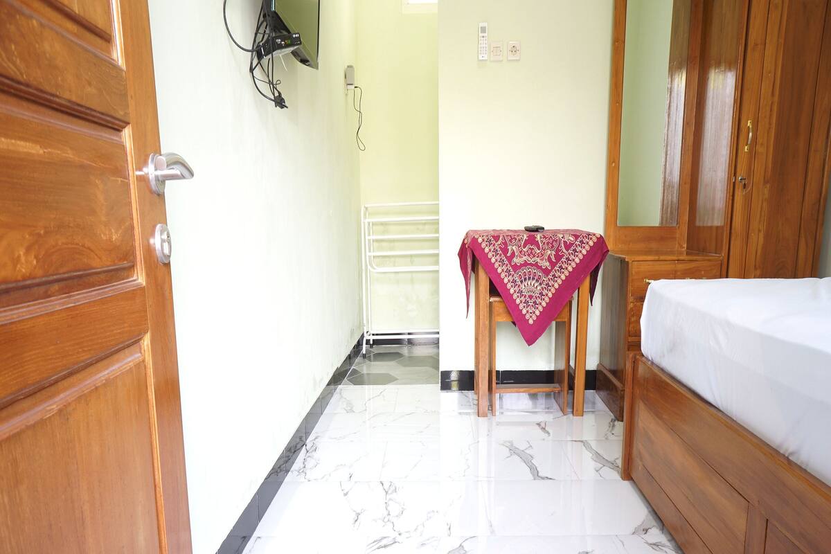 A hotel room with a wooden wardrobe, a bed with a white mattress, a marble floor, and a small table with a red tablecloth.