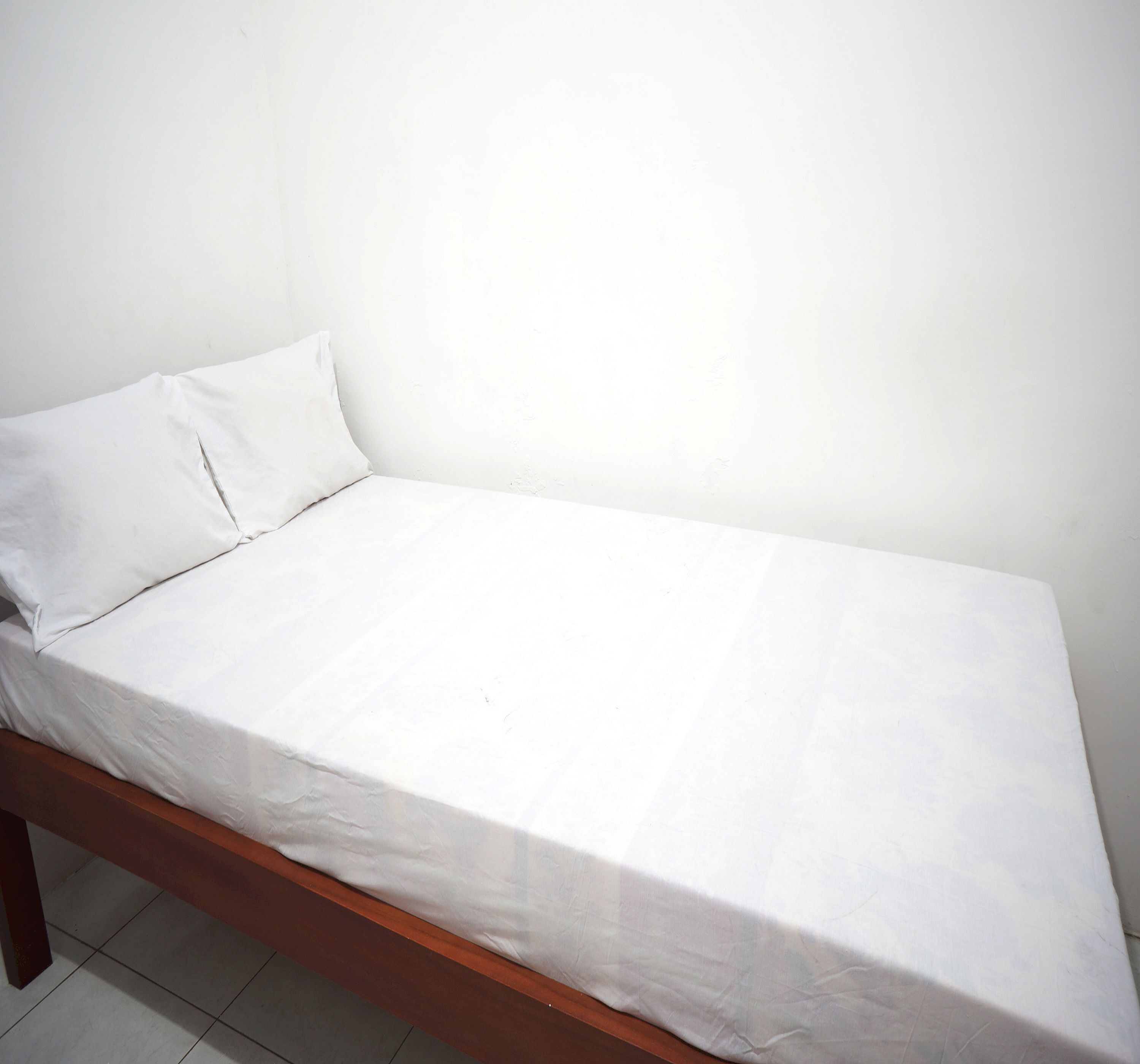 A single bed with a wooden frame and white bedding in a room with plain white walls and tiled flooring.