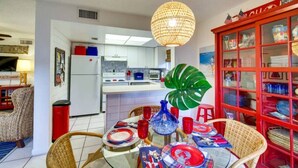 A modern kitchen with white cabinets, a dining area with wicker chairs, a red cabinet with glassware, and a large green plant. - Beachside Cottage - 5 min walk to the Beach (Cocoa Beach)