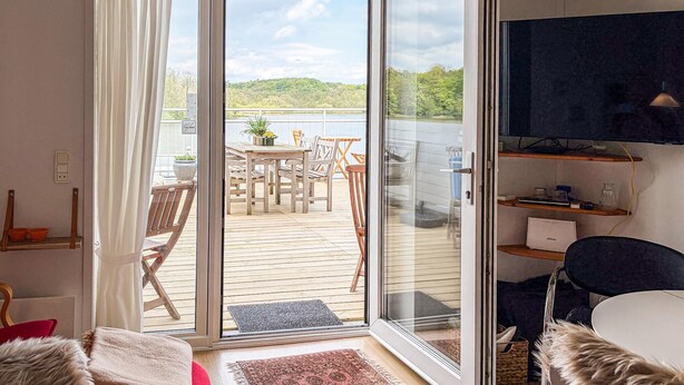 Ein Zimmer mit einer Schiebeglastür, die auf eine Terrasse mit Tisch und Stühlen führt, einem Fernseher auf einem Regal und einer Felldecke auf dem Bett.