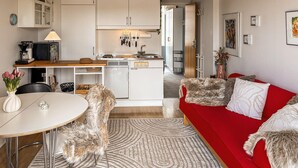 Eine moderne Küche mit weißen Schränken, einem Einbauofen und einer Spüle. Ein Essbereich mit rundem Tisch und Stühlen, sowie ein rotes Sofa mit Fellkissen.