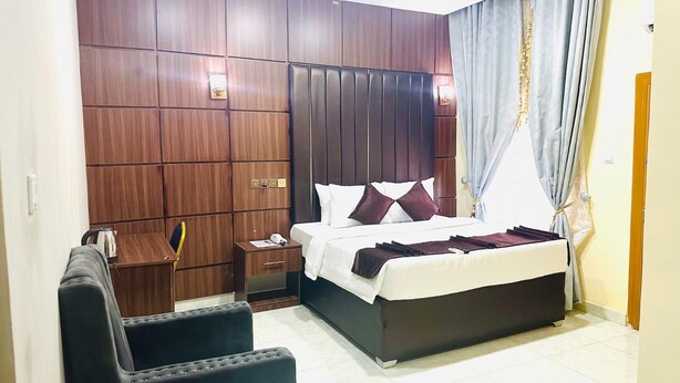 A hotel room with a large bed, a wooden wardrobe, a small table with a kettle, and a chair. - Davina hill resorts and hotel  (Abuja)