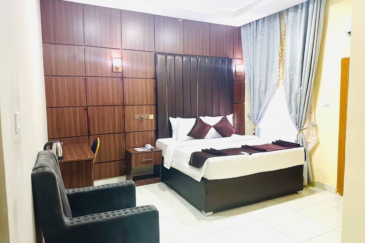 A hotel room with a large bed, a wooden wardrobe, a small table with a kettle, and a chair.
