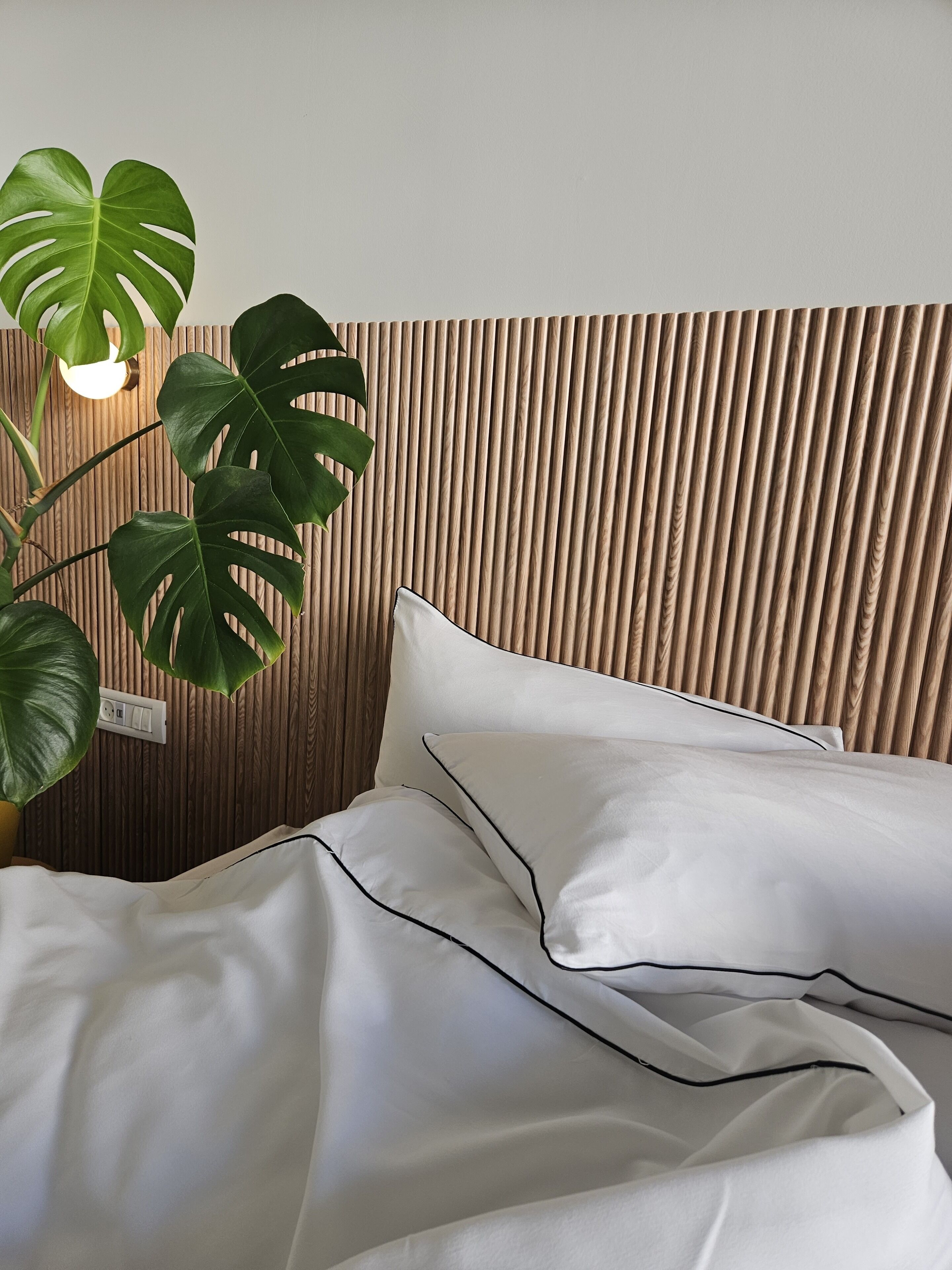 Una cama con ropa de cama blanca y detalles negros, un cabecero de listones de madera y una planta verde con una lámpara colgante.