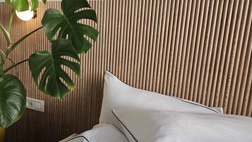 Una cama con ropa de cama blanca y detalles negros, un cabecero de listones de madera y una planta verde con una lámpara colgante.