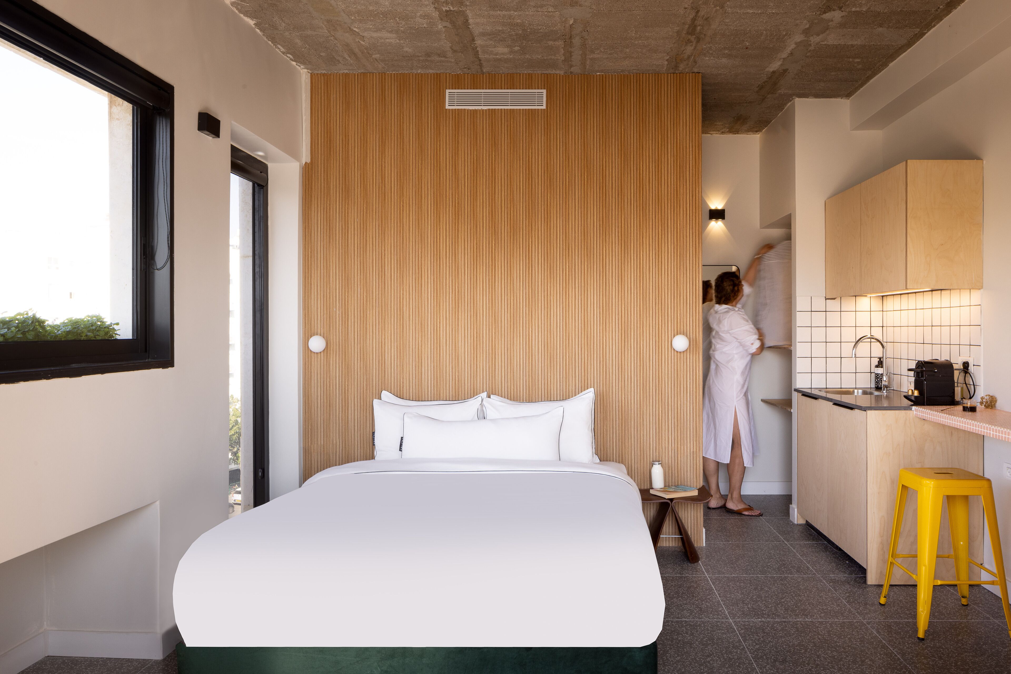 A modern hotel room with a large bed, a wooden accent wall, a kitchen area, and a yellow stool.