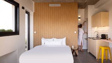 A modern hotel room with a large bed, a wooden accent wall, a kitchen area, and a yellow stool.