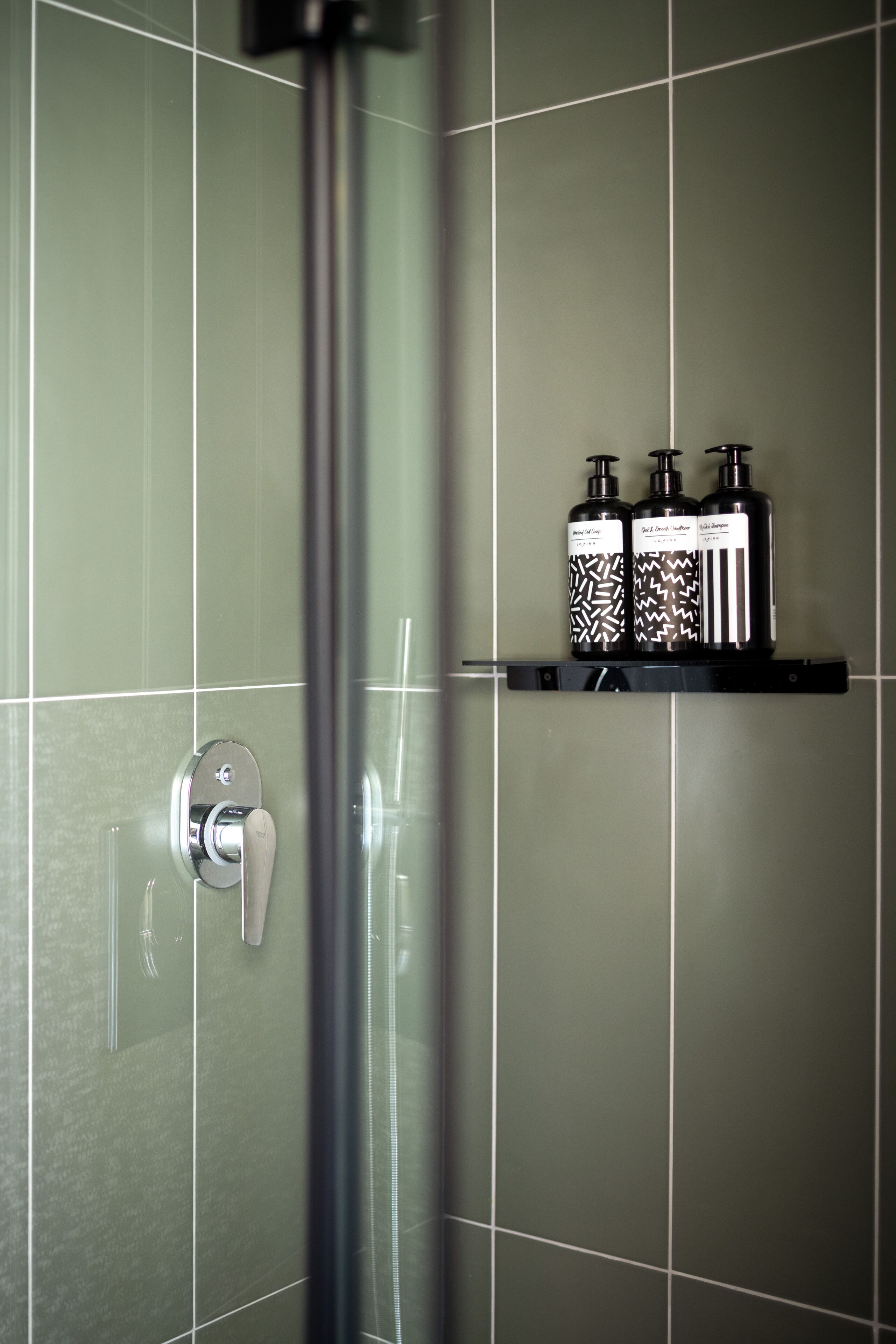 Un baño con azulejos verdes, una llave de ducha y tres botellas de artículos de aseo en un estante.