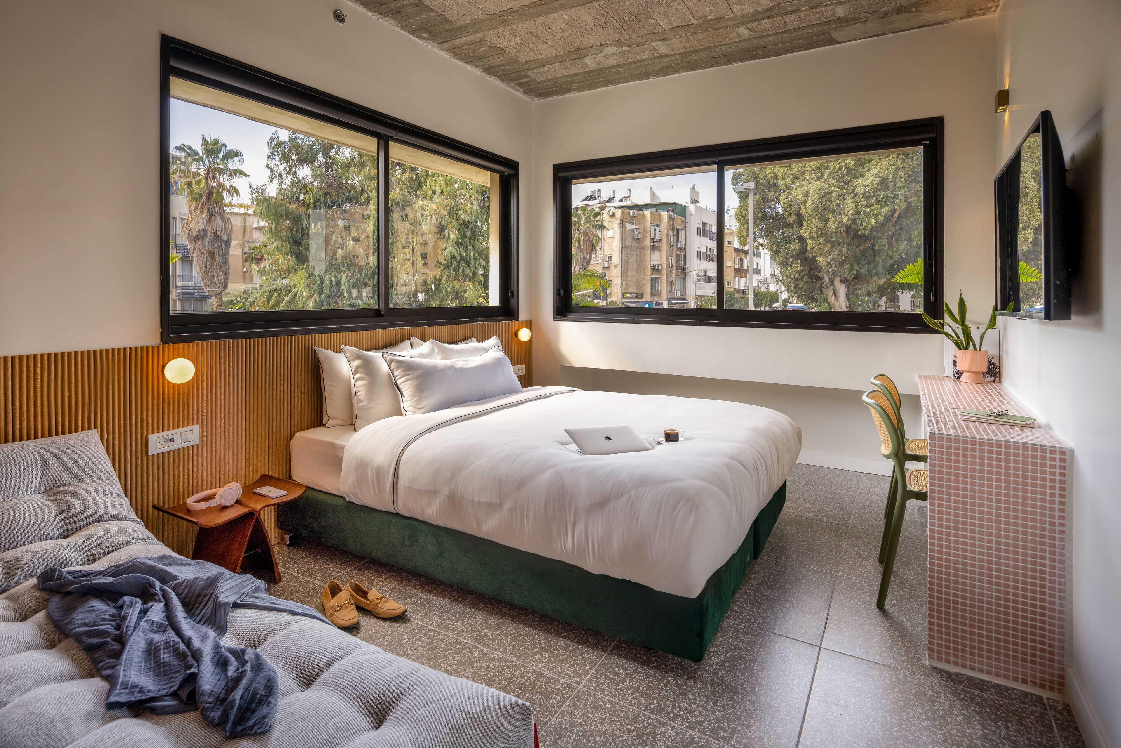 A bedroom with a bed, bedside table, chair, and a view of buildings and trees through large windows.