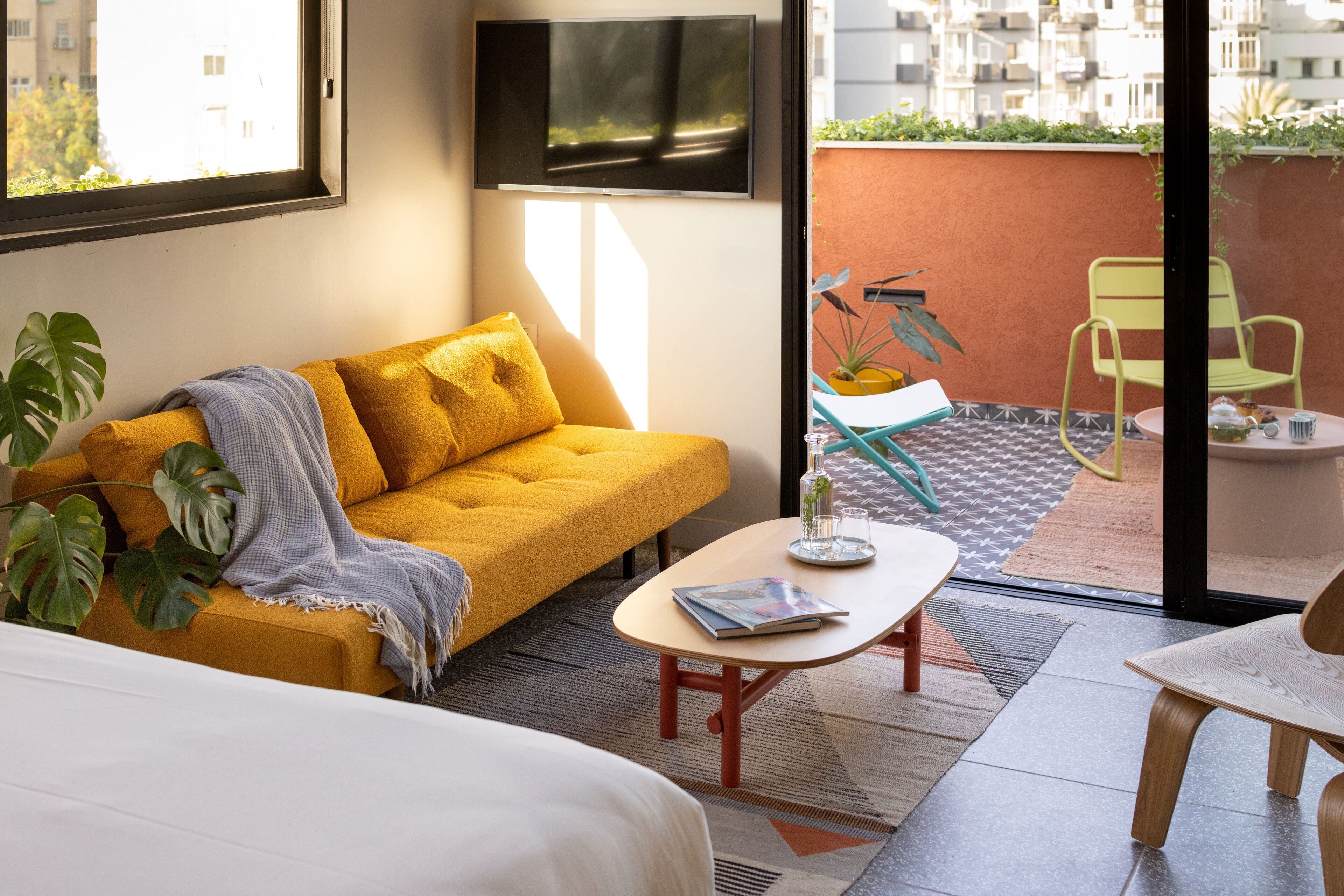 A modern hotel room with a yellow sofa, a small table with a magazine, a large window, and a view of the outside balcony area.