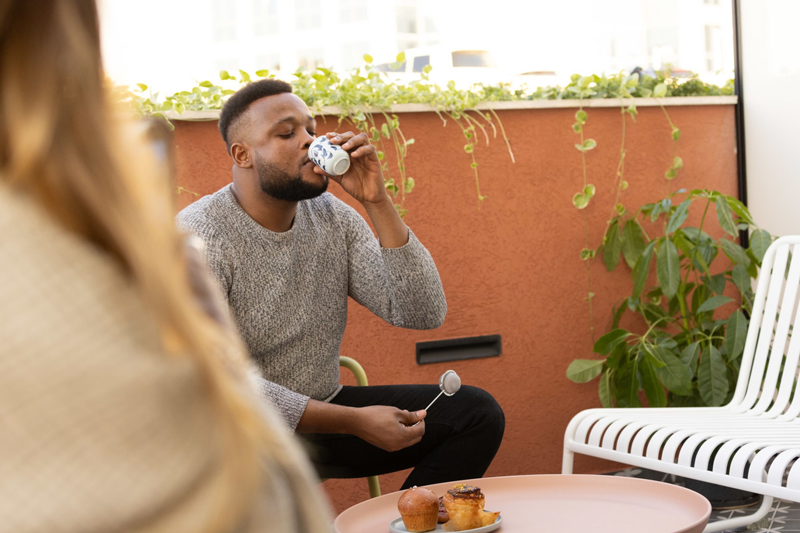 A person sitting at a table with a cup and a muffin, with a white chair and a plant in the background.
