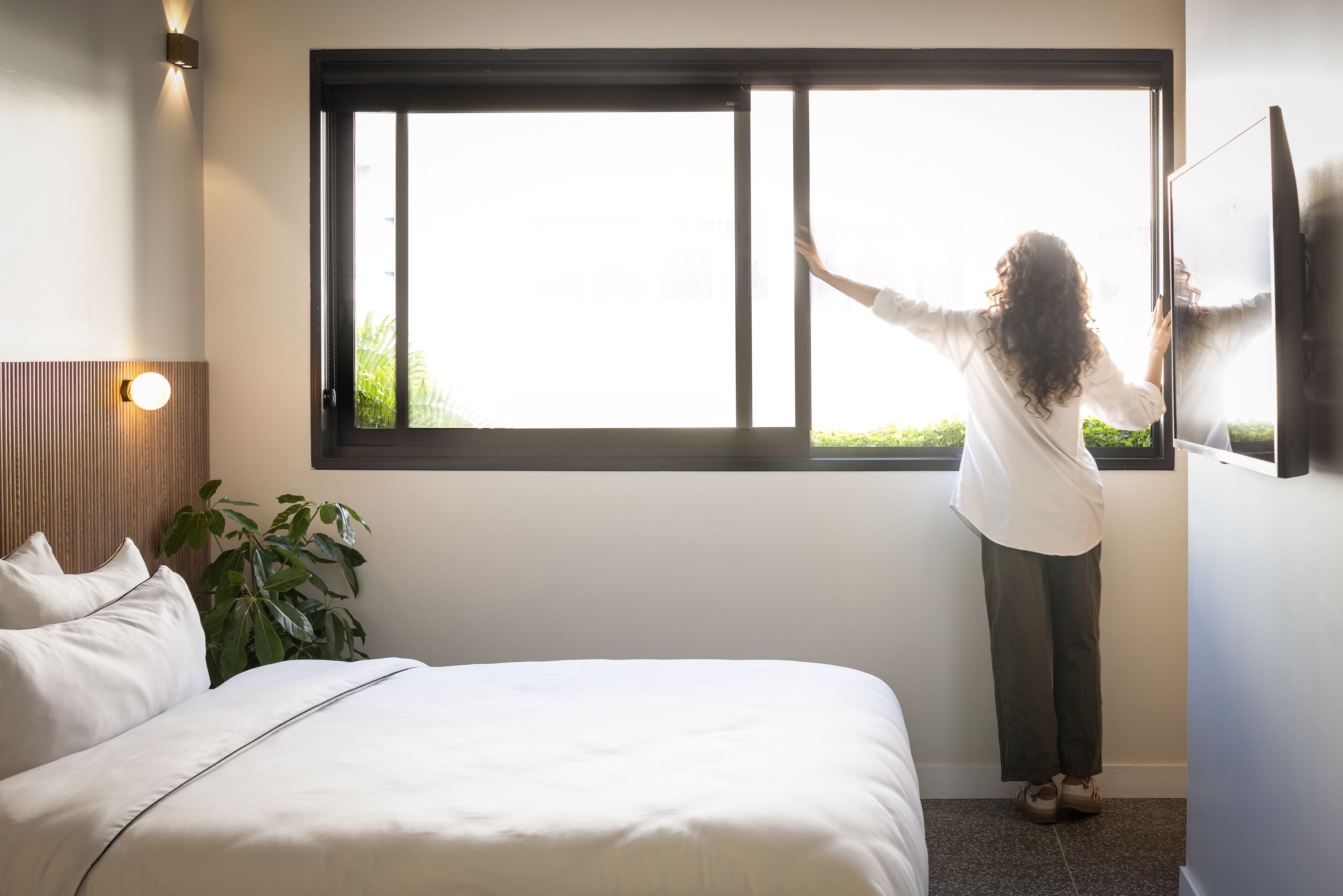 Una persona de pie junto a una ventana en una habitación con una cama y una planta.
