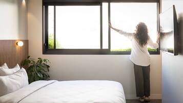 Una persona de pie junto a una ventana en una habitación con una cama y una planta.
