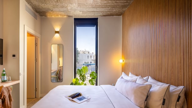A hotel room with a large bed, a bedside table with a bottle and glasses, a mirror, and a window with a view of buildings. - Savanna Dizengoff - Smart Hotel by Loginn Tel Aviv (Tel Aviv)