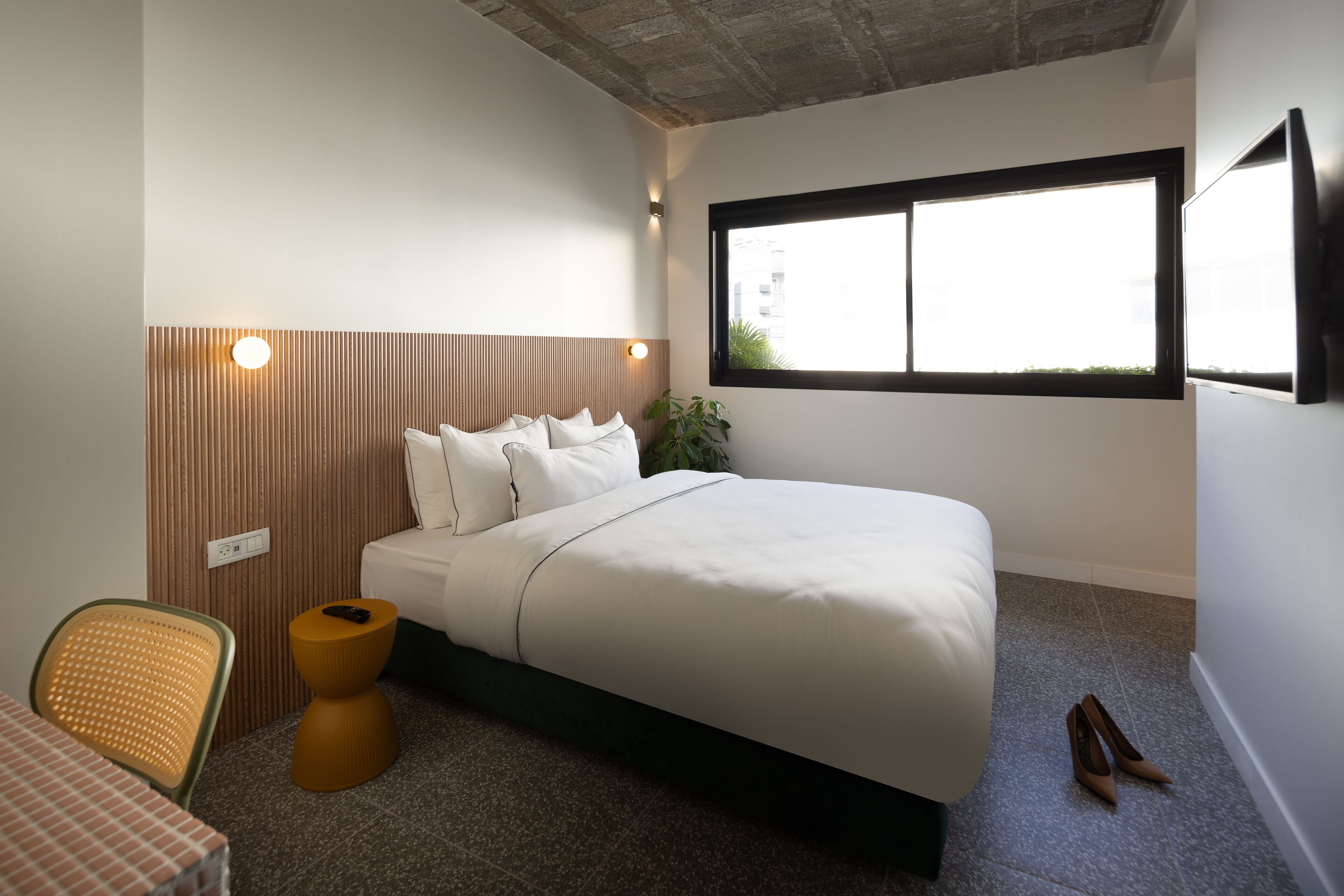 A bedroom with a bed, a chair, a small table, a window, and a plant.