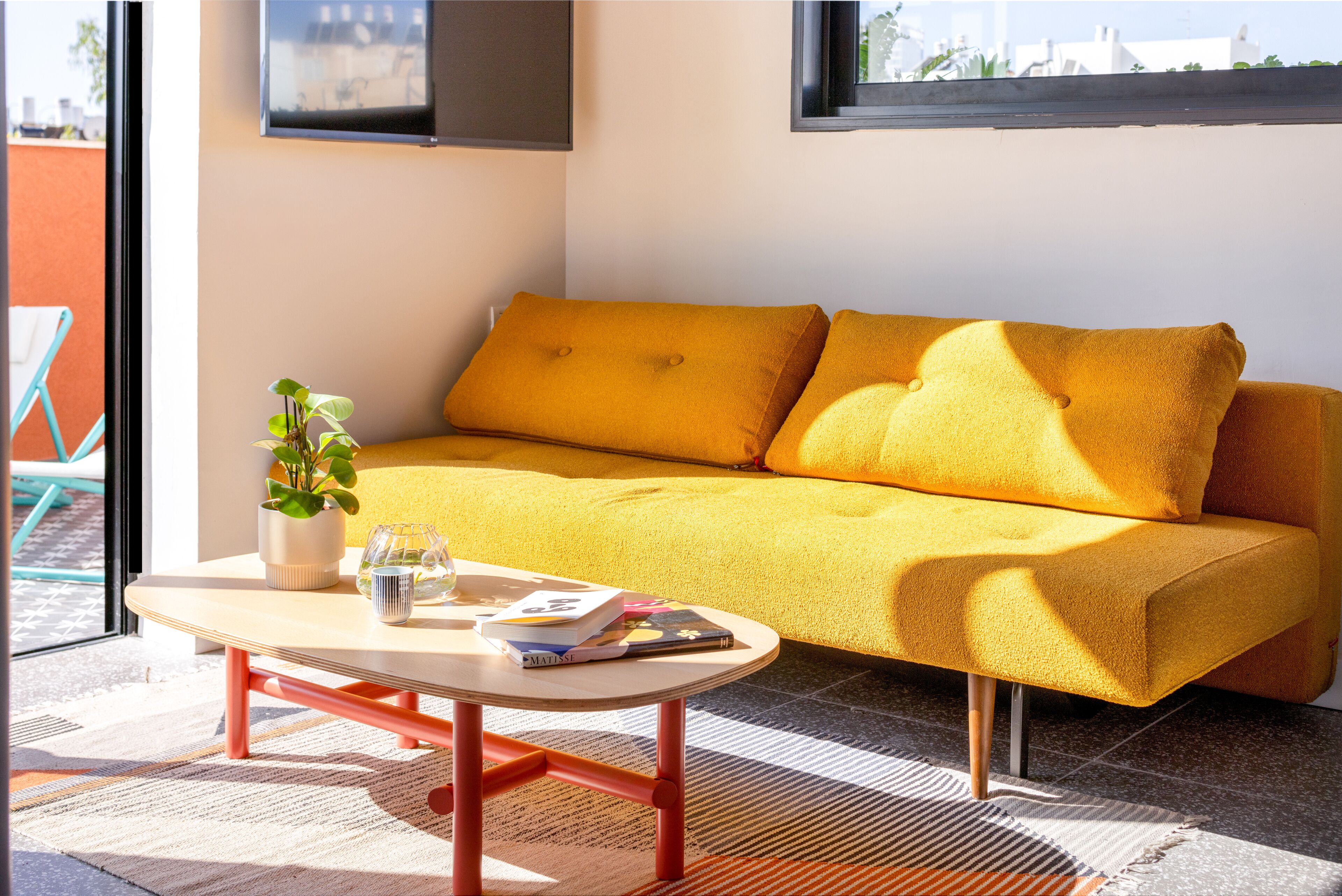 A modern living room with a yellow sofa, a small table with books and a plant, and a view of the city through a window.