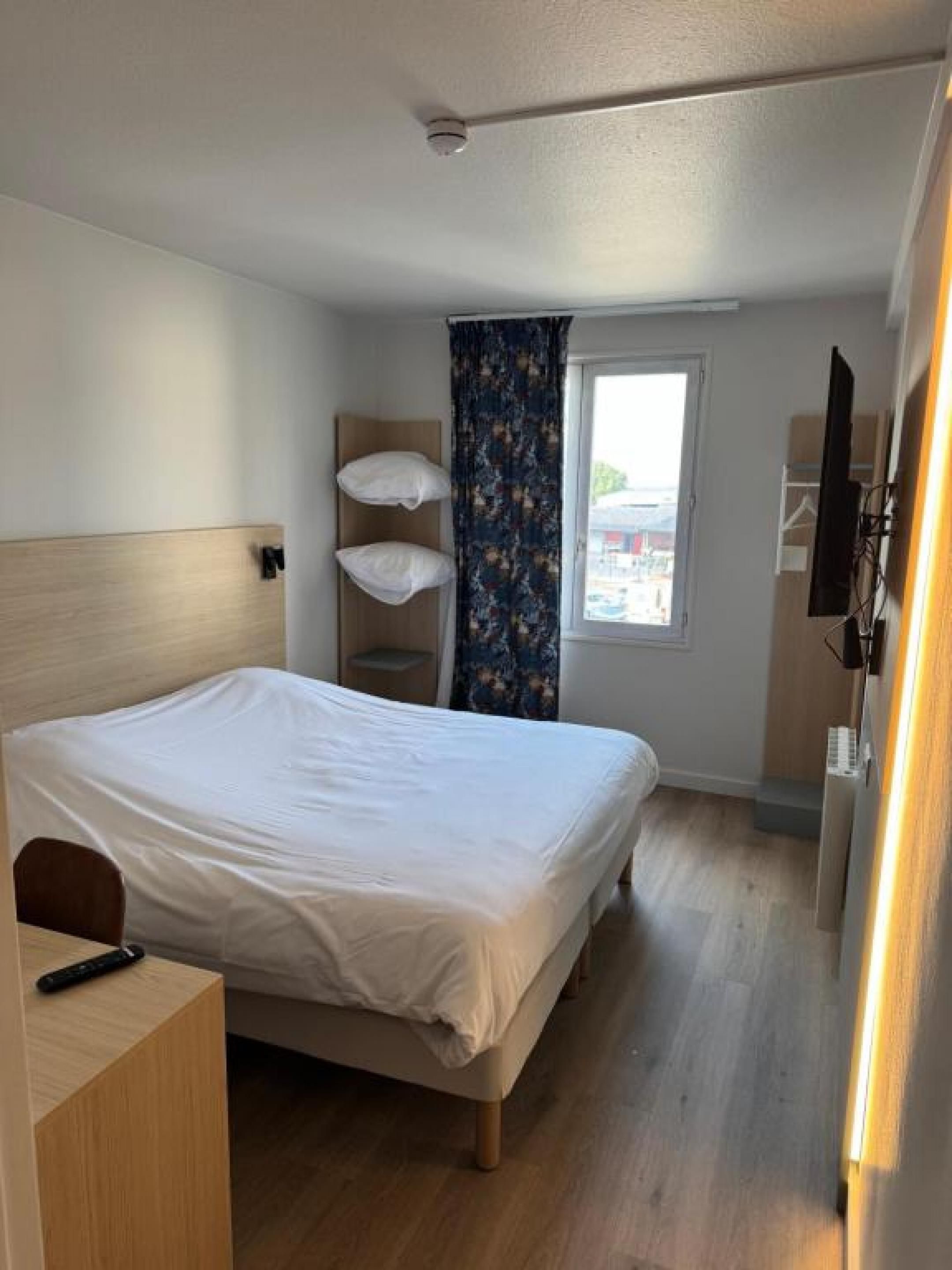 Una habitación de hotel con cama, escritorio, televisor y ventana con cortinas.