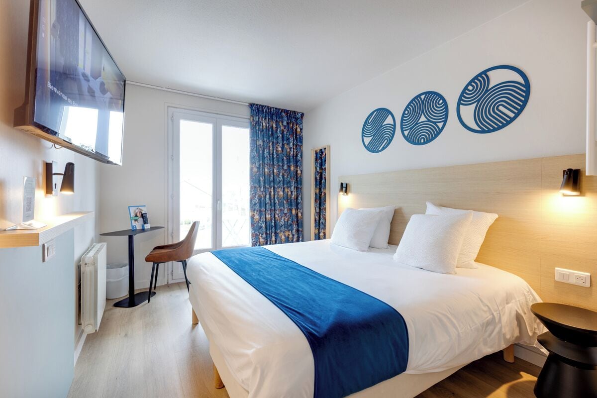 Quarto de hotel com cama, escrivaninha, cadeira, televisão e janela com cortinas.