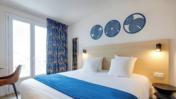 A hotel room with a large bed, a wooden headboard, a side table, and a chair. - Brit Hotel Confort Carhaix (Carhaix-Plouguer)