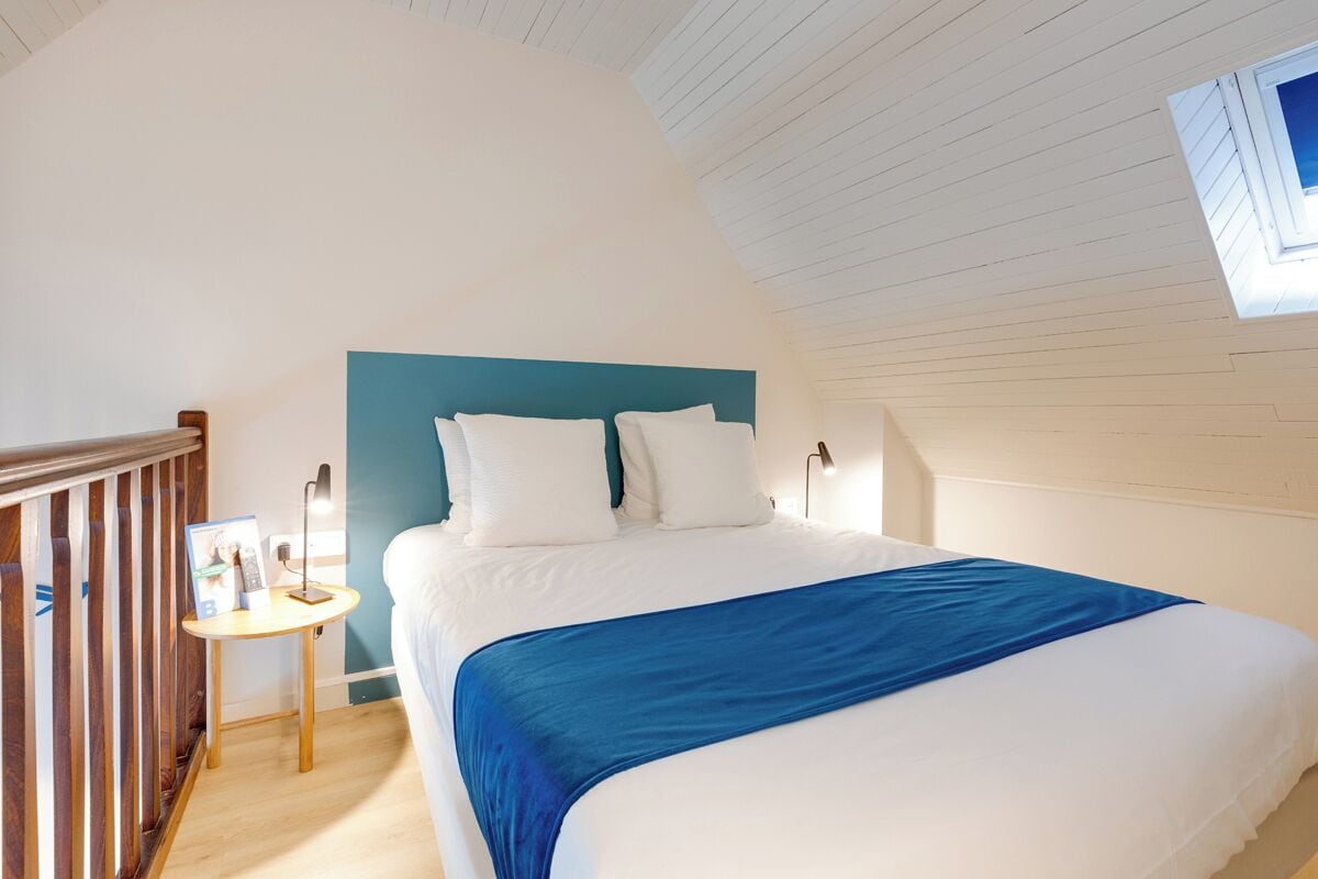 A bedroom with a bed, a bedside table, a nightstand, a window, and a sloped ceiling.