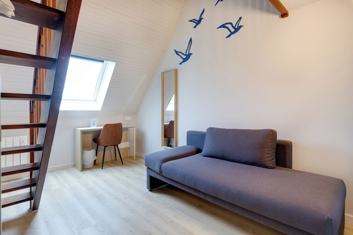 Una habitación tipo loft con techo inclinado, un escritorio integrado, un sofá, una silla y una escalera.