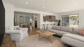 A spacious living area with a sofa, a coffee table, and a TV. There is a kitchen in the background with a dining area.