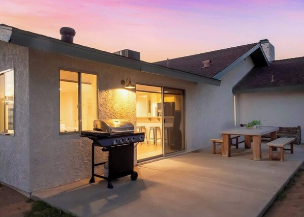 A modern house with a patio, a barbecue grill, and a dining area with a table and benches. - Stardust Ranch | Hot tub + Double living rooms (Yucca Valley)