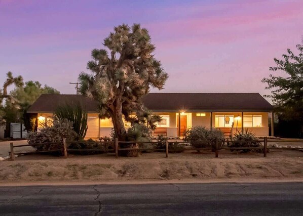 A two-story house with a desert landscape, a large tree in front, and a well-lit front porch.