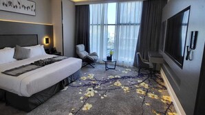 A modern hotel room with a large bed, a seating area with a chair and a small table, a flat-screen TV mounted on the wall, and a large window with sheer curtains. - msharef almoden hotel (Riyadh)