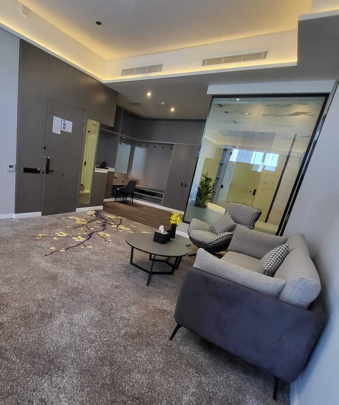 A modern living room with a sofa, armchair, and coffee table. There is a sliding glass door leading to a kitchen area.