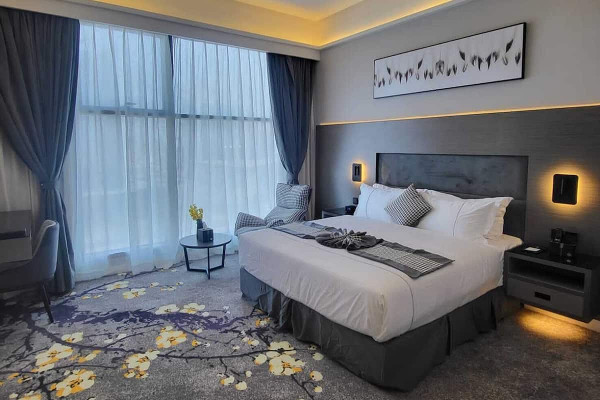 A modern hotel room with a large bed, a small side table, a chair, and a large window with curtains.