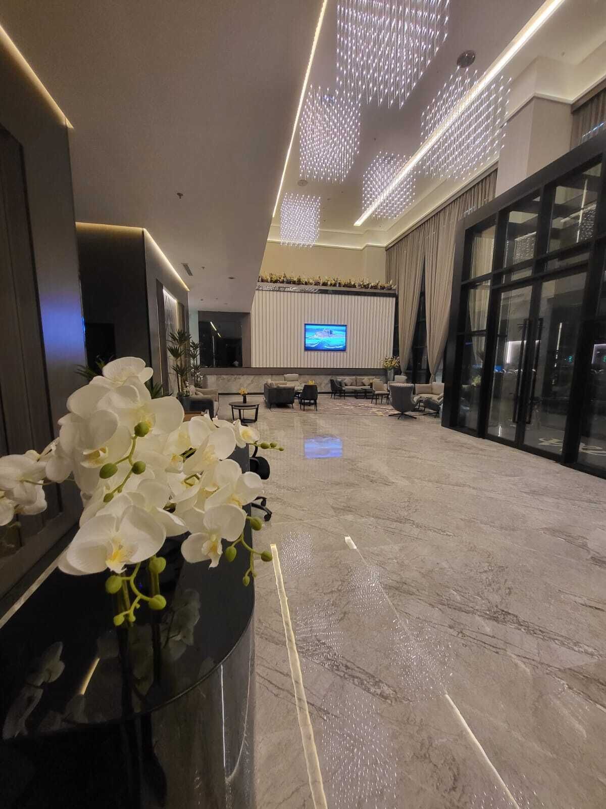 A spacious lobby with a marble floor, a reception desk with white orchids, a large flat-screen TV, and modern lighting fixtures.