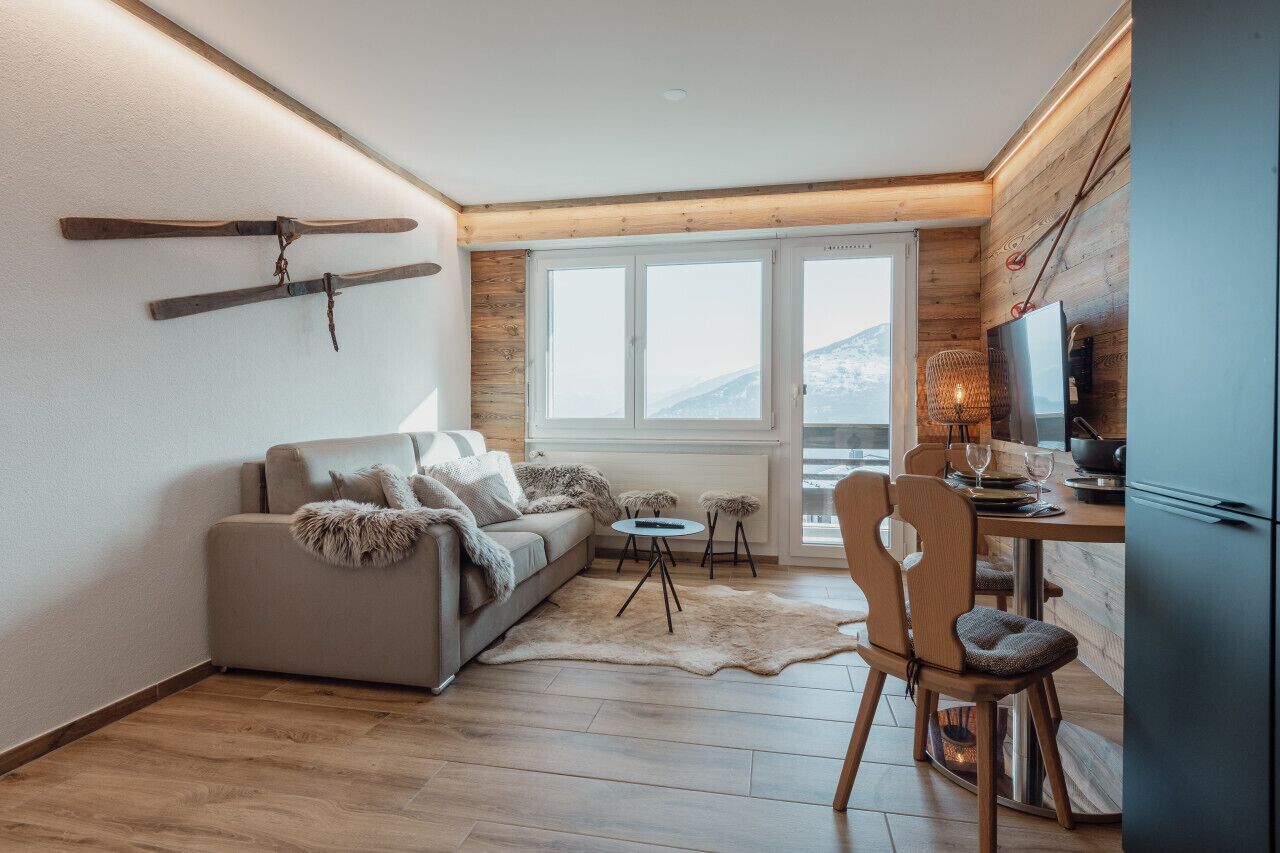 Un salón acogedor con sofá, mesa de comedor y televisión. Hay ventanas con vistas a montañas.