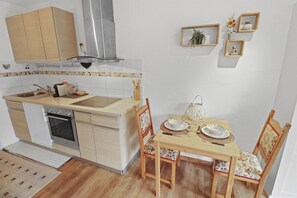 A compact kitchen with a sink, stove, and cabinets, a dining table with chairs, and a wall-mounted shelf with decorative items.