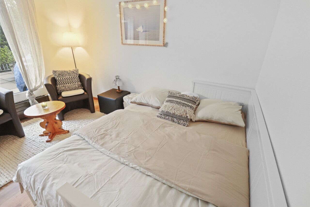 Un dormitorio con una cama, una silla, una mesa, una lámpara y un cuadro en la pared.