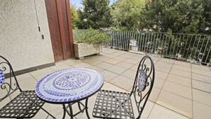 A patio with a mosaic table and metal chairs, surrounded by a metal fence and trees. - Charmant Studio Ouvert Avec vue Paisible (Crans-Montana)