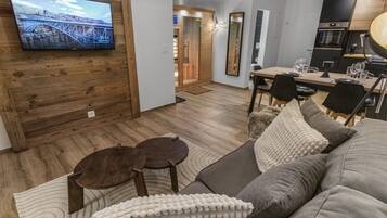Ein modernes Wohnzimmer mit einer Wandvertäfelung aus Holz, einem Flachbildfernseher, einem Essbereich mit Holztisch und -stühlen sowie einer Küchenzeile mit Herd und Kühlschrank.