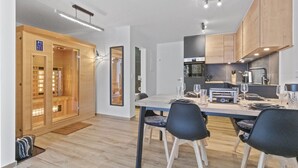 A modern kitchen with wooden cabinets, a dining table set for four, and a sauna visible through a glass door. - Appartement rénové avec vue panoramique et sauna (Saas-Fee)
