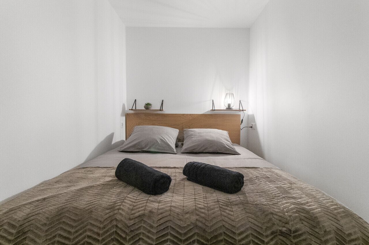 A small, narrow hotel room with a single bed, two pillows, a wooden headboard, a bedside table with a lamp, and two rolled towels on the bed.