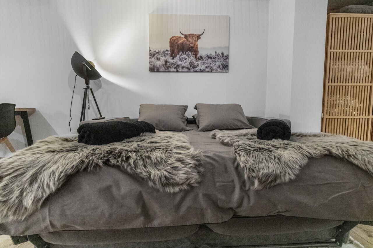 Een slaapkamer met een bed, twee kussens, een bont plaid, een staande lamp en een ingelijste foto aan de muur.