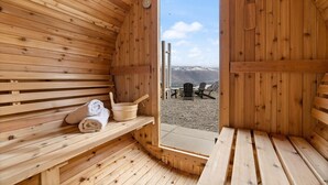 Un sauna en bois avec des bancs et une banquette, une autre banquette en bois avec des serviettes, et une vue sur une plage et des montagnes à l’extérieur.