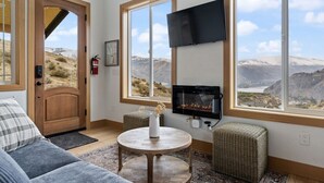 Une pièce chaleureuse avec une cheminée, un canapé, une télévision et de grandes fenêtres donnant sur une vue imprenable sur les montagnes.