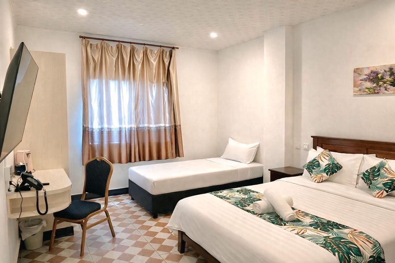 A hotel room with a checkered floor, a desk with a telephone, a chair, two beds with different bedding, and a window with curtains.